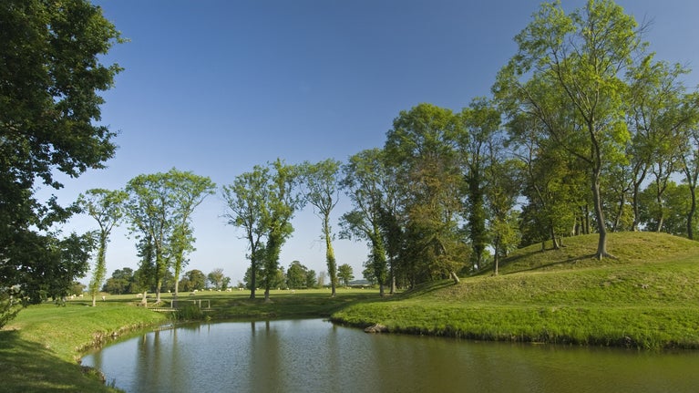 A canal curves round a spiral mound, up the mound and on the banks of the canal are large trees in full leaf.